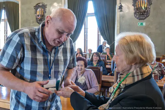 Mężczyzna wręcza medal starszej kobiecie w sali z innymi uczestnikami.