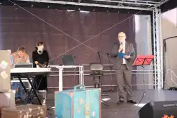 Mężczyzna w garniturze przemawia do publiczności, obok dwie osoby przy stole.