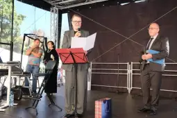 Kobieta przemawia do publiczności, obok niej mężczyzna z dokumentami, w tle inni uczestnicy wydarzenia.