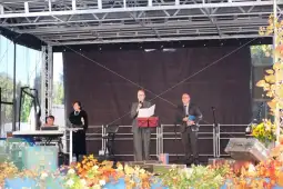 Mężczyzna w garniturze przemawia na scenie, obok niego stoi druga osoba, w tle instrumenty muzyczne.