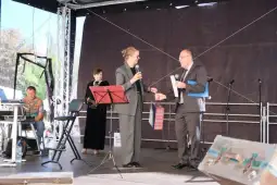 Dwóch mężczyzn rozmawia na scenie, obok muzycy grają na instrumentach.