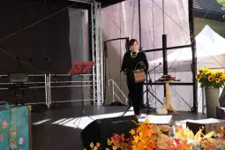 Kobieta na scenie z torbą, w otoczeniu mikrofonów i dekoracji.