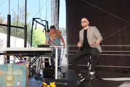 Mężczyzna w okularach przeciwsłonecznych siedzi na krześle, obok muzyk przy keyboardzie.