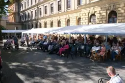 Grupa osób siedzących na zewnątrz pod namiotem, w tle budynek.