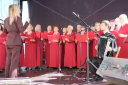 Chór kobiet w czerwonych sukienkach śpiewa pod kierunkiem dyrygenta, akompaniament na akordeonie.