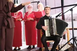 Mężczyzna gra na akordeonie, w tle chór kobiet w czerwonych strojach.