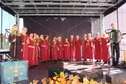 Chór w czerwonych strojach z kwiatami na scenie, grający z akordeonem.