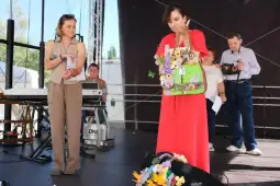 Kobieta w czerwonej sukience trzyma torbę z kwiatami, obok druga kobieta z mikrofonem.