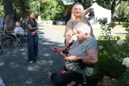 Kobieta z mikrofonem przemawia, a w tle grupa osób, w tym starsza kobieta na wózku.