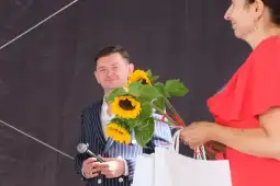 Mężczyzna w marynarce z mikrofonem i kobieta w czerwonej sukience trzymająca bukiet słoneczników.