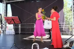 Kobieta w różowej sukience wręcza kwiaty drugiej kobiecie w czerwonej sukience na scenie.