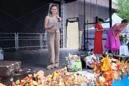 Kobieta na scenie trzymająca kartkę, obok inne osoby w kolorowych strojach. Dekoracje z liści.