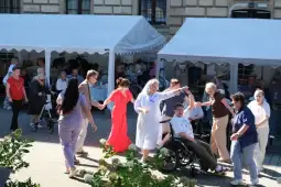 Seniorzy tańczą na świeżym powietrzu, w tle namioty i wózki inwalidzkie.
