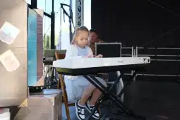 Dziewczynka gra na pianinie na scenie, w tle mężczyzna przy komputerze.
