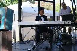 Chłopiec gra na pianinie, obok stoi dorosła osoba przy sprzęcie muzycznym.