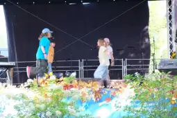 Dzieci tańczą na scenie, w tle mikrofony i dekoracje roślinne.