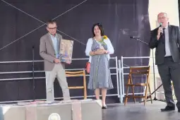 Dwóch mężczyzn i kobieta na scenie, jeden trzyma obraz, kobieta ma słonecznik.