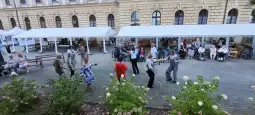 Grupa ludzi tańczących na świeżym powietrzu w otoczeniu namiotów i krzewów.