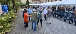 Grupa osób tańczących w kręgu na zewnątrz, w otoczeniu namiotów i krzeseł.