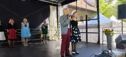 Mężczyzna i kobieta w eleganckich strojach występują na scenie, w tle inne osoby.
