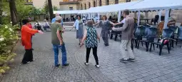 Grupa osób tańczących na zewnątrz, w otoczeniu krzeseł i zieleni.