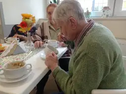 Starsza osoba pije z filiżanki, w tle inne osoby przy stole z jedzeniem i kwiatami.