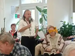 Mężczyzna z mikrofonem prowadzi zajęcia dla grupy osób starszych. Kobieta z opaską na oczach bierze udział w zabawie.