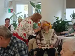 Kobieta z mikrofonem rozmawia z osobą z zawiązanymi oczami w otoczeniu innych seniorów.