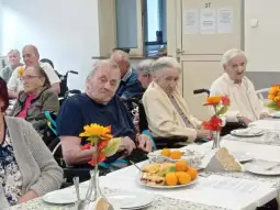 Starsze osoby siedzą przy stole z jedzeniem i kwiatami, w tle widać innych seniorów.