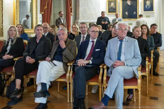 Grupa osób siedzących w eleganckim wnętrzu, uczestniczących w wydarzeniu.