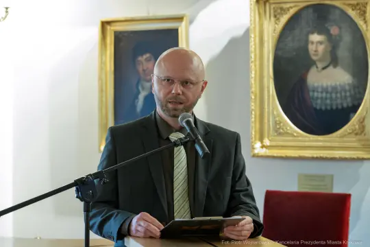 Mężczyzna przemawia przy mikrofonie w sali z obrazami w tle.