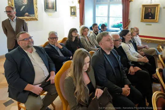 Grupa ludzi siedzących w sali, w tle obrazy i okna.
