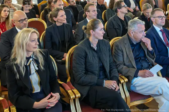 Grupa ludzi siedzących na wydarzeniu, w eleganckich strojach, z poważnymi wyrazami twarzy.