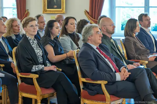 Grupa osób siedzących na krzesłach w sali, skupionych na wydarzeniu.