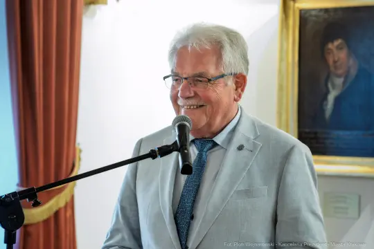 Mężczyzna w jasnym garniturze uśmiecha się i przemawia do mikrofonu.