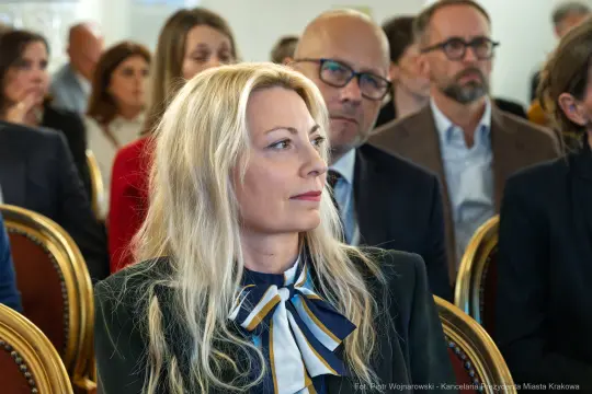 Kobieta z długimi blond włosami w eleganckim stroju, słuchająca wystąpienia w tłumie.