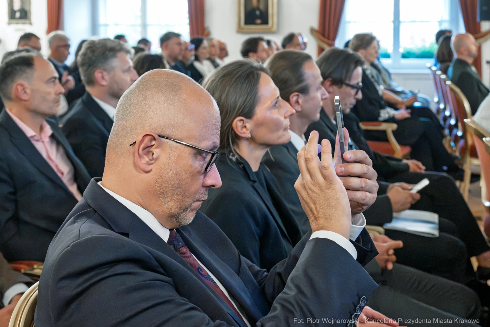 Grupa ludzi w formalnych strojach, niektórzy z telefonami, uczestniczy w wydarzeniu.  Autor: Piotr Wojnarowski