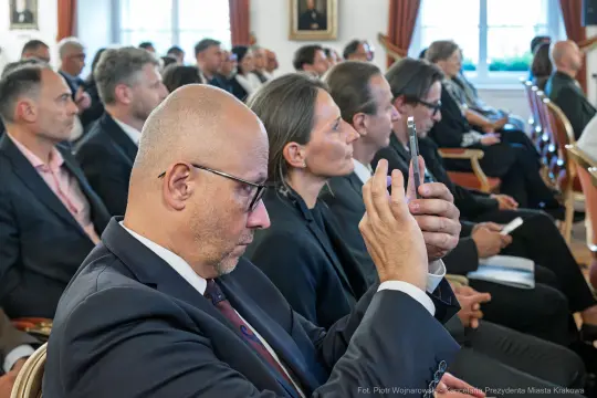 Grupa ludzi w formalnych strojach, niektórzy z telefonami, uczestniczy w wydarzeniu.