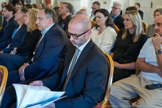 Grupa ludzi siedzących na sali, niektórzy trzymają dokumenty.