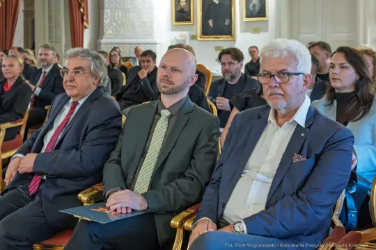Trzech mężczyzn w garniturach siedzi na sali wśród innych uczestników wydarzenia.