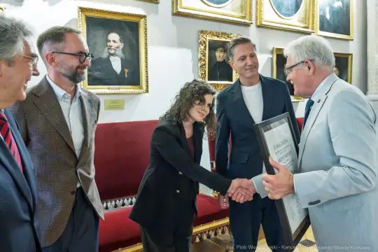Wręczenie nagrody w towarzystwie kilku osób w eleganckim wnętrzu.