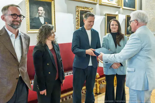 Wręczenie nagrody w uroczystej atmosferze, cztery osoby w formalnych strojach.