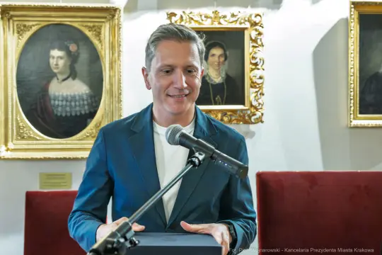 Mężczyzna w garniturze trzyma mikrofon, stoi przed obrazami w eleganckim wnętrzu.