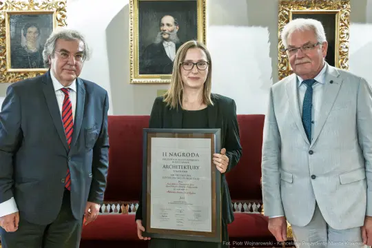 Kobieta trzymająca dyplom nagrody architektonicznej, obok dwóch mężczyzn w garniturach.