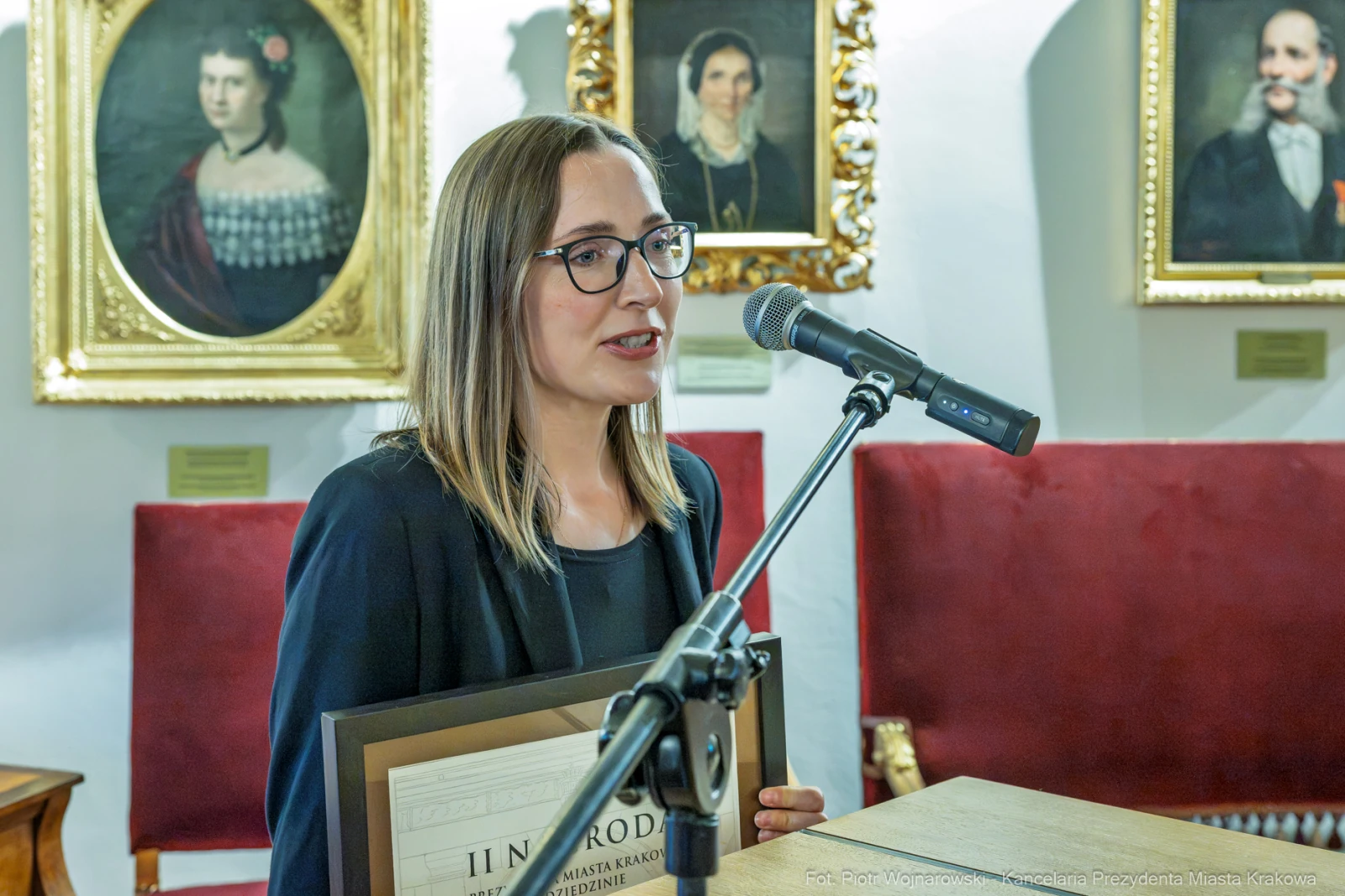Kobieta przemawia przy mikrofonie, trzymając dyplom, w tle portrety na ścianie.  Autor: Piotr Wojnarowski