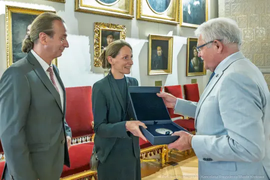 Wręczenie nagrody w eleganckim wnętrzu z portretami na ścianach.