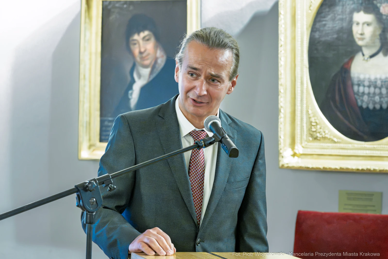 Mężczyzna w garniturze przemawia przy mikrofonie, w tle portrety.  Autor: Piotr Wojnarowski