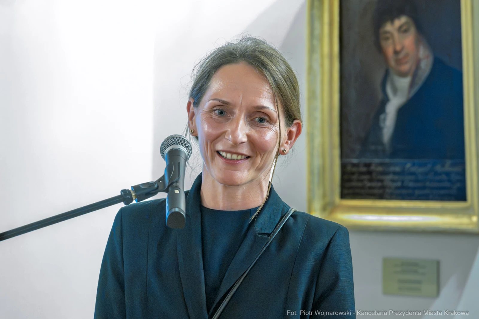 Kobieta uśmiechająca się podczas wystąpienia publicznego, w tle portret.  Autor: Piotr Wojnarowski