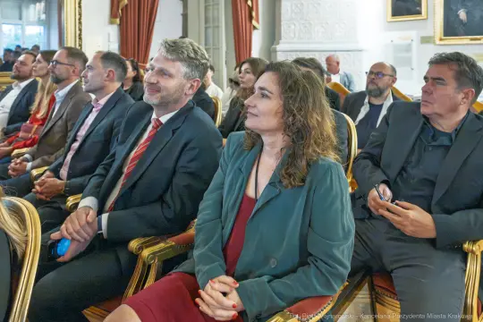 Grupa osób siedzących w eleganckiej sali, uczestniczących w wydarzeniu.