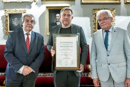 Mężczyzna trzymający dyplom nagrody architektonicznej, obok dwóch mężczyzn w garniturach.